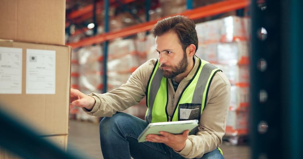 Funcionário conferindo mercadorias com tablet em depósito, mostrando a rotina prática do controle de estoque e organização de produtos.