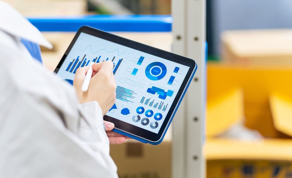 Profissional analisando gráficos de controle em tablet dentro de um armazém, representando o monitoramento de dados e a gestão estratégica dos diferentes tipos de estoque.