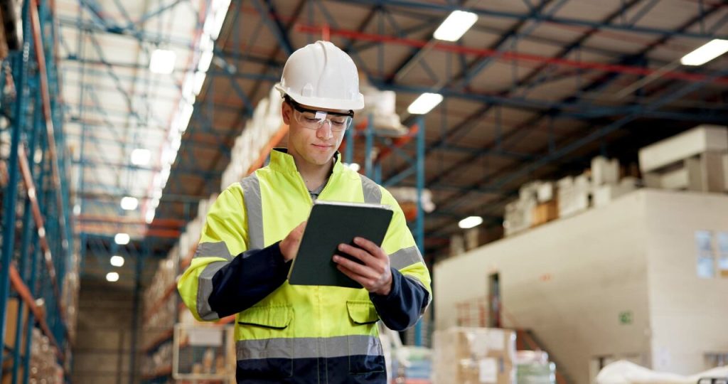 Profissional utilizando tablet para gestão de estoque em armazém logístico, com prateleiras organizadas e operação de supply chain ao fundo