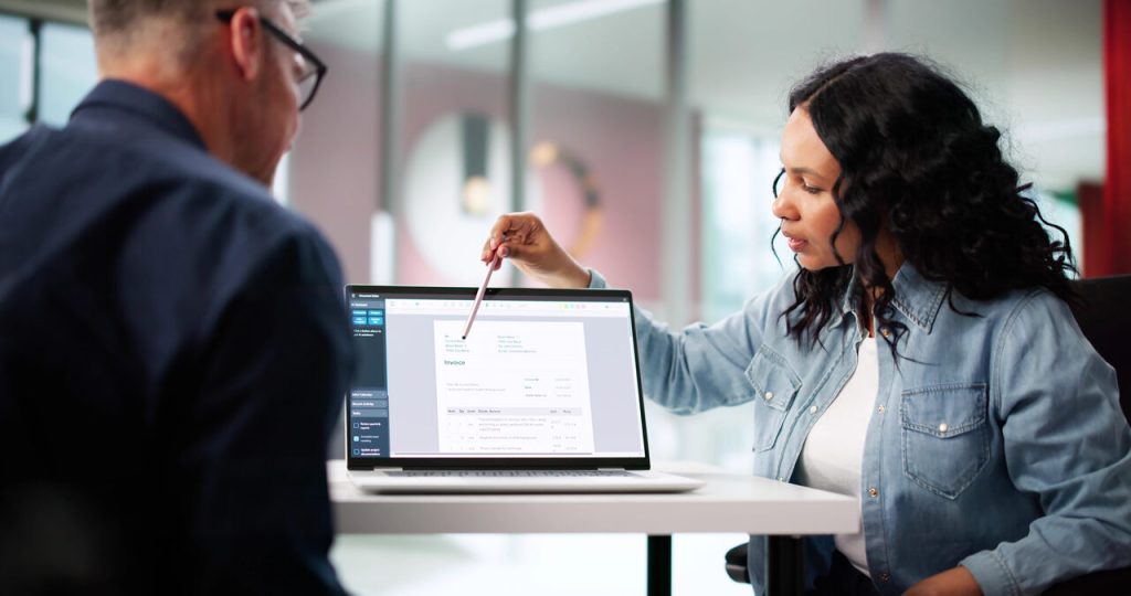 Mulher apresentando declaração de prestação de serviços em laptop para colega em ambiente corporativo, destacando informações do documento na tela.