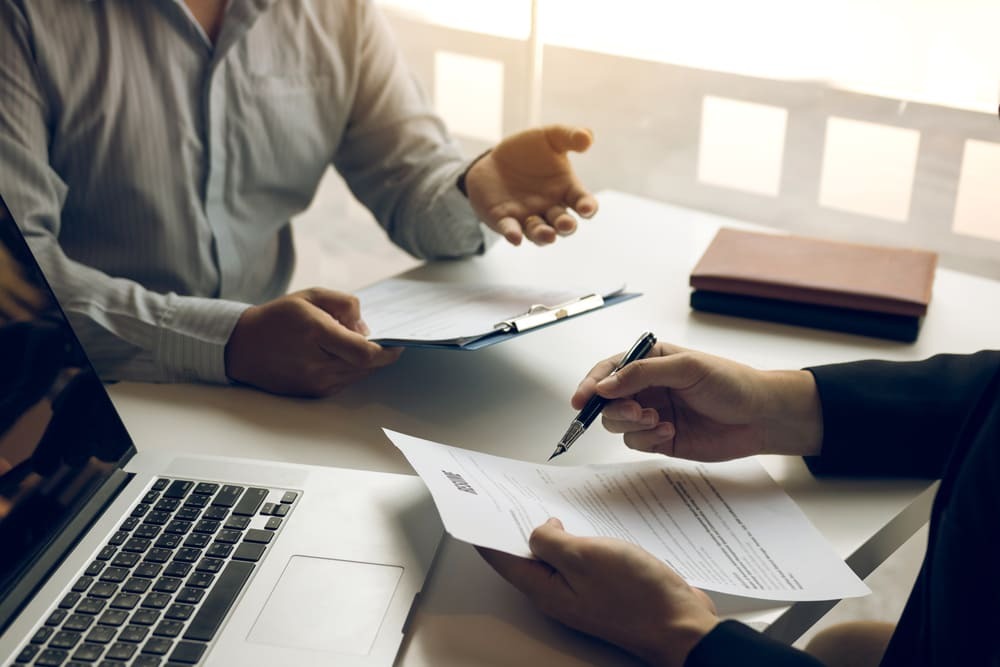 Profissionais analisando e assinando minuta de contrato durante reunião corporativa com documentos e notebook sobre a mesa.