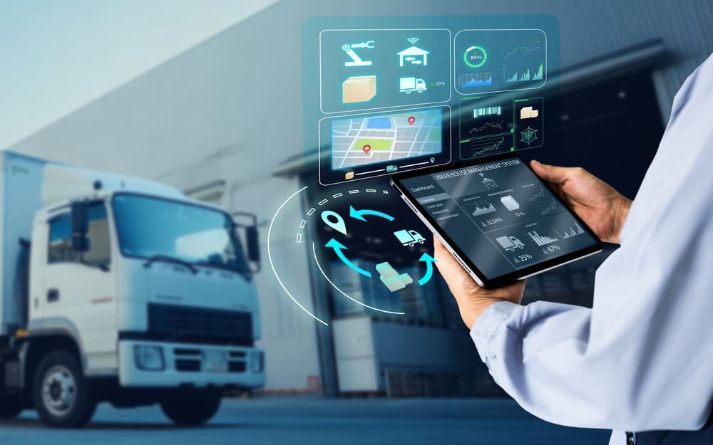 Homem utilizando tablet para monitorar operações e indicadores dos tipos de logística, com caminhão ao fundo em ambiente de armazém representando transporte e gestão da cadeia de suprimentos.