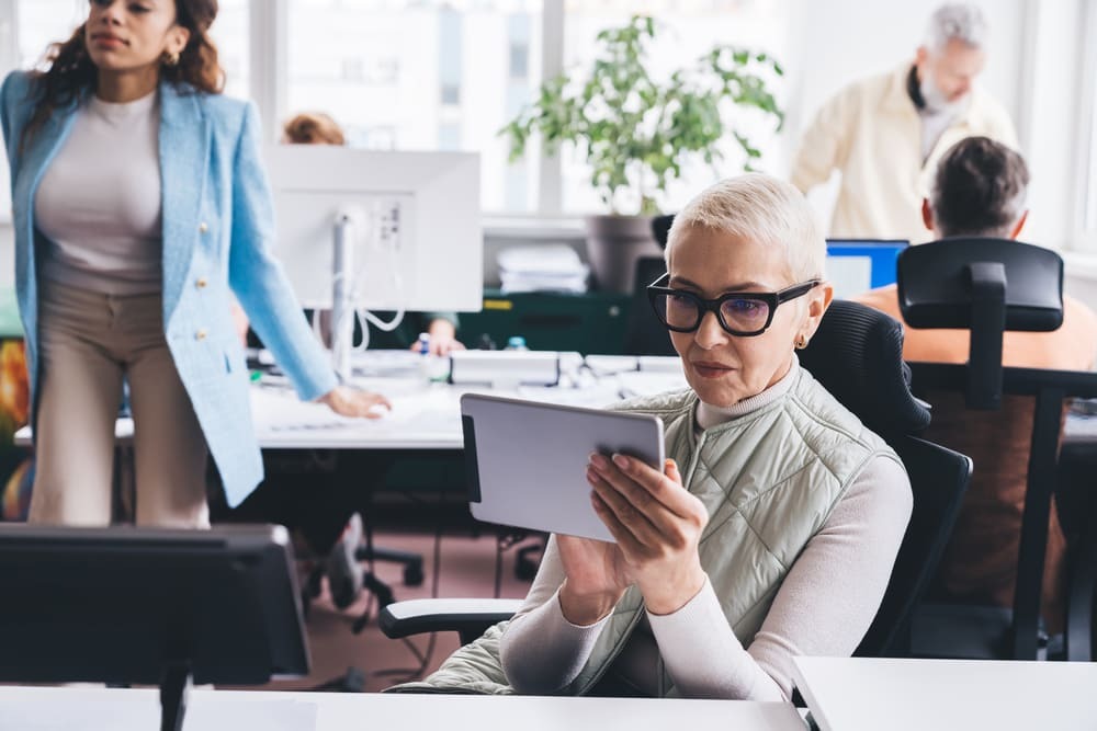 Mulher de cabelos curtos grisalhos sentada em cadeira no escritório, segurando tablet em mãos enquanto utiliza sistema de controle de ponto para registro de jornada.