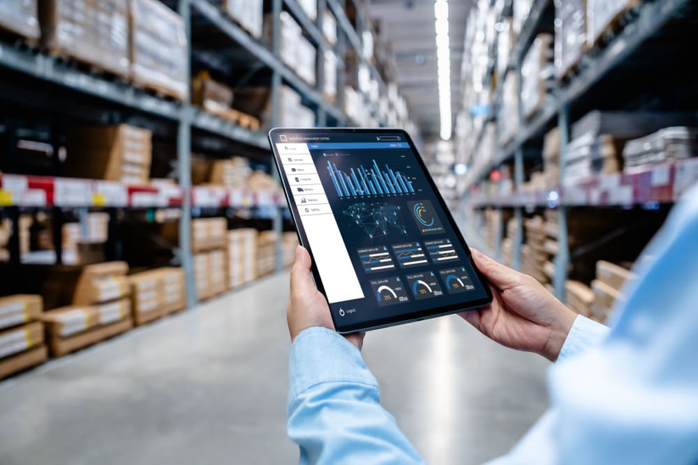 Gestor analisando dados na logística em um tablet dentro de um centro de distribuição, com dashboards de desempenho, estoque e movimentação de mercadorias.