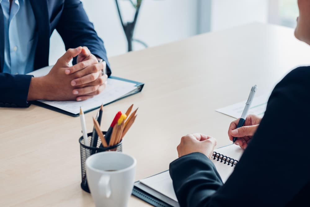 Foco nas mãos de líder conversando com colaborador em reunião de feedback corretivo, anotando ações de melhoria em caderno para acompanhar evolução.