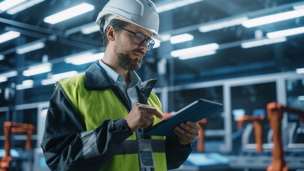 Técnico industrial com capacete e tablet realiza controle e acompanhamento de processos da produção industrial em um chão de fábrica moderno.