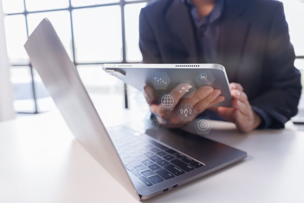 Executivo utilizando notebook e tablet com ícones de tecnologia, ilustrando a integração de sistemas e o acesso às informações de gestão em diferentes dispositivos.