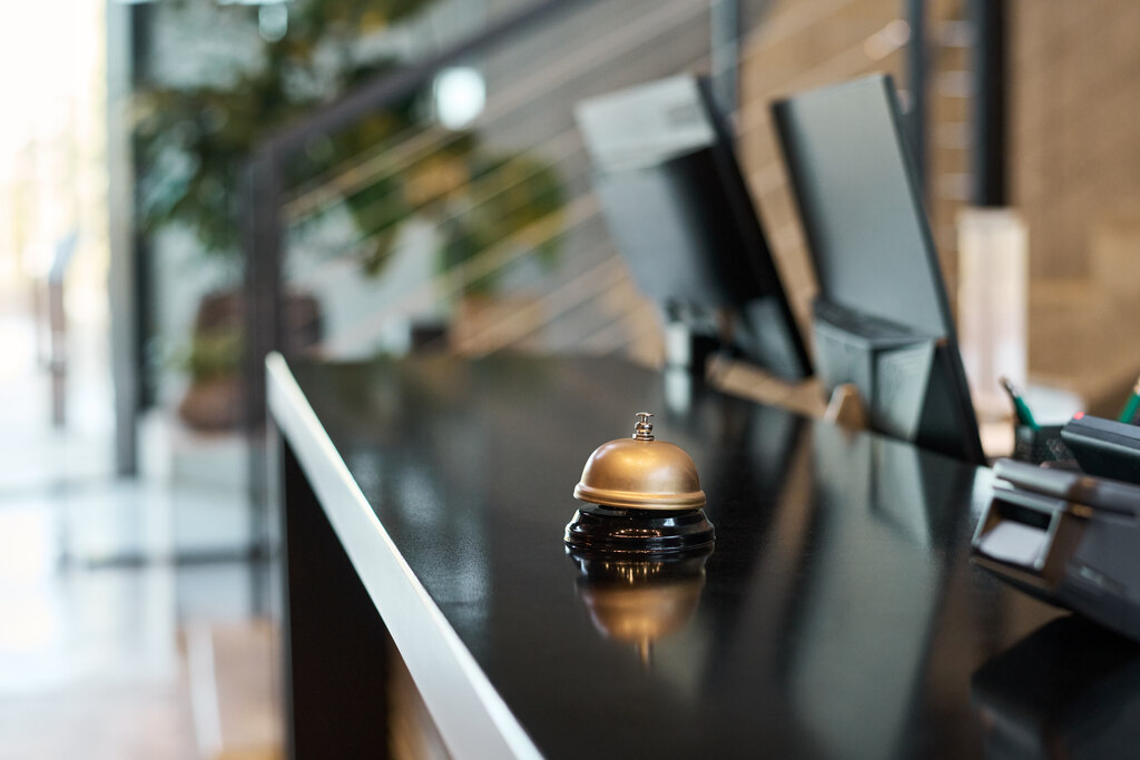 Fotografia em plano detalhe de um balcão de recepção moderno com uma sineta dourada em destaque sobre a superfície preta brilhante, simbolizando a transição entre o atendimento clássico e o uso de tecnologias para check-in e check-out em hotéis. Ao fundo, de forma levemente desfocada, aparecem monitores de computador e terminais de autoatendimento que agilizam o registro e a saída dos hóspedes, integrando a tradição da hospitalidade com a inovação digital.