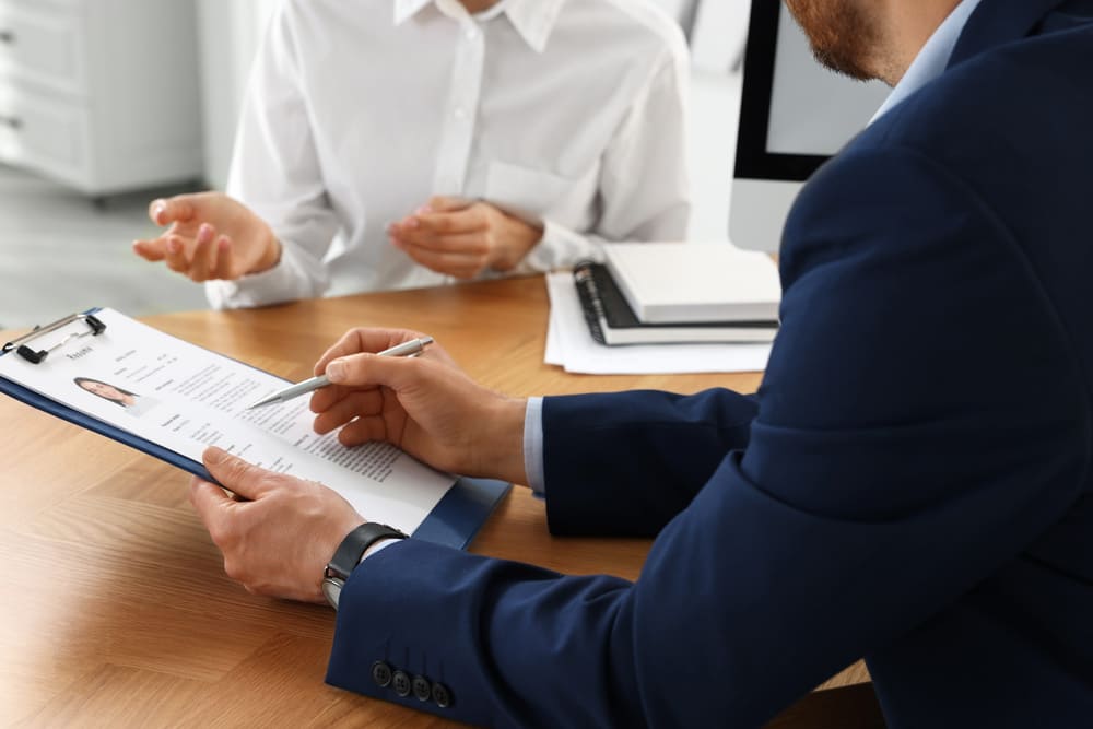 Entrevistador analisando o currículo enquanto conversa com a candidata, representando exemplos do que falar na entrevista de emprego e como apresentar experiências profissionais.