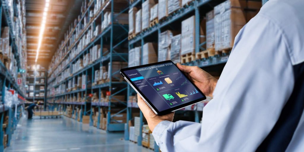 Sistema de gestão logística em armazém com operador analisando dados em tablet e prateleiras organizadas ao fundo
