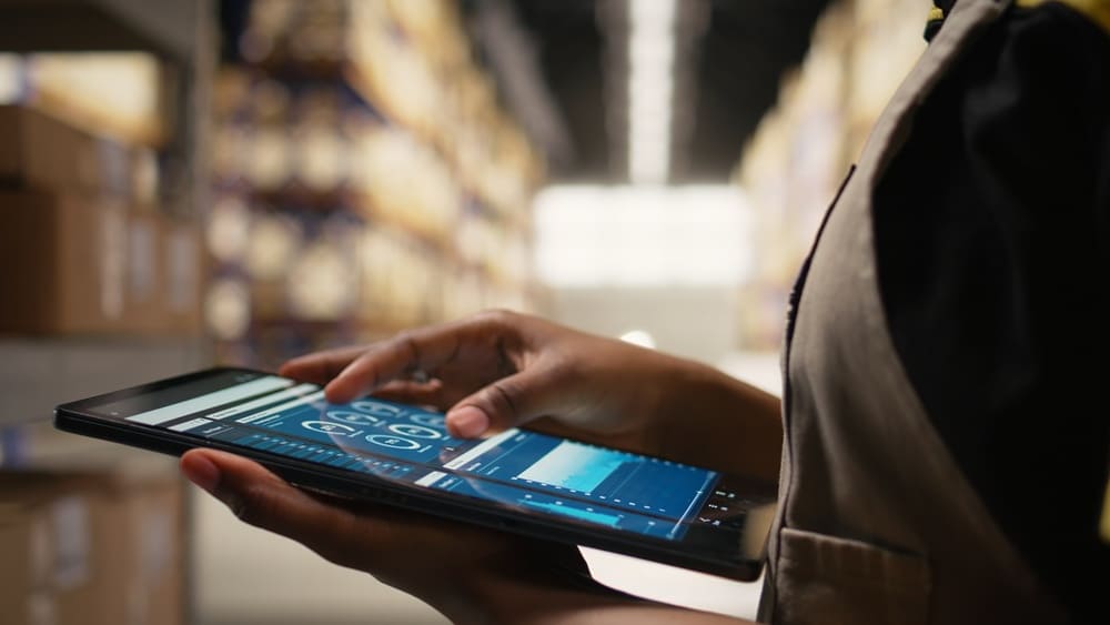 Mulher mexendo em tablet com dados sobre expedição de mercadorias. O foco está nas mãos da mulher e, ao fundo, está o interior de um armazém desfocado.