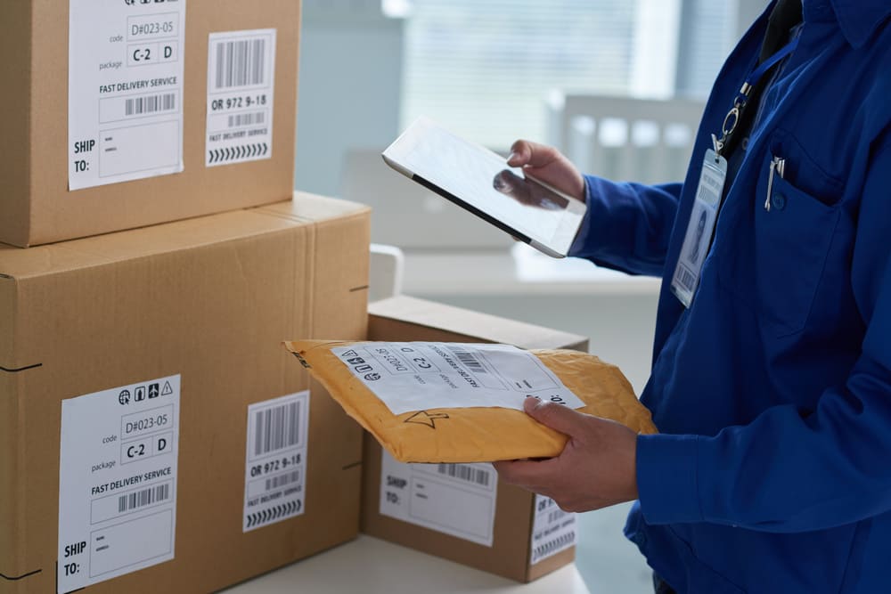 Homem vestido com uma camisa azul, segurando um tablet em uma mão e um pacote amarelo na outra, em frente a caixas enquanto organiza o envio de mercadorias para empresas de logística.
