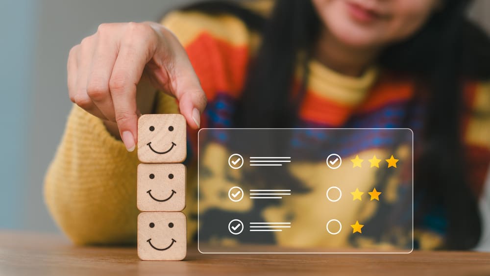 Mulher com blusa colorida empilha blocos com rostos sorridentes ao lado de avaliação com estrelas, representando análise de satisfação e gestão de riscos psicossociais nas empresas.