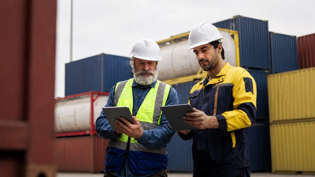 Profissionais de logística utilizam tablets em área portuária com contêineres, representando a gestão de operações de comércio exterior e Global Trade Management