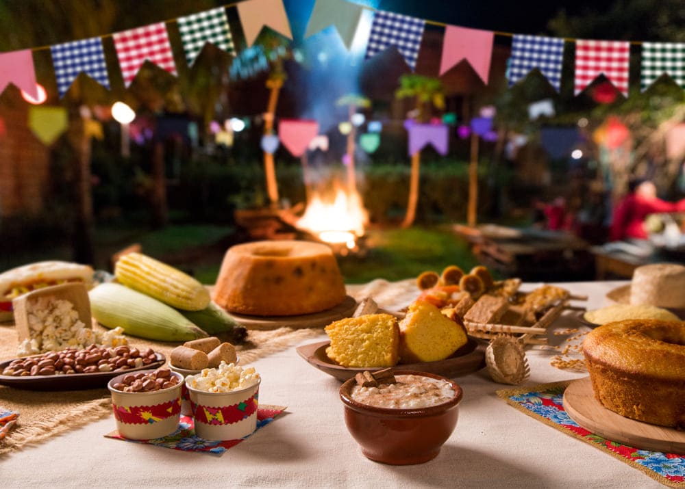 Mesa com comidas típicas para festa junina na empresa, como cuscuz, pipoca, milho, bolos e canjica. Uma fogueira está ao fundo da mesa bandeirinhas aparecem acima e ao fundo da imagem.